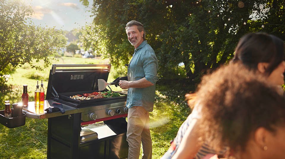 Hombre cocinando en una barbacoa Weber de gas en un jardín con su familia esperando al fondo.